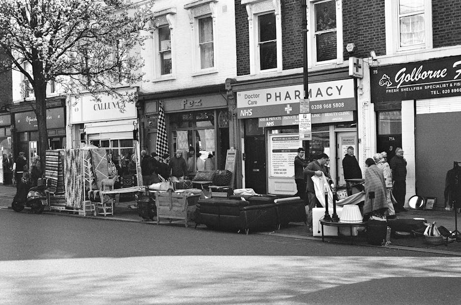 A black and white photograph of a busy urban street scene showing a row of small shops, including a pharmacy and a fish and seafood specialist store. Outside the shops, various household items and furniture, such as sofas, chairs, and a lamp, are arranged on the pavement, likely for collection or sale. Several pedestrians are walking past or viewing the items, with some engaged in conversation. A small scooter is parked on the left side of the image, close to the curb. The surroundings include brick building facades and tree branches partially visible at the top left. The overall scene suggests an area where private rubbish or bulk waste is being temporarily displayed for removal or collection, aligning with alternative waste handling services like those offered by Rubbish Removal Wimbledon, near Wimbledon Broadway.