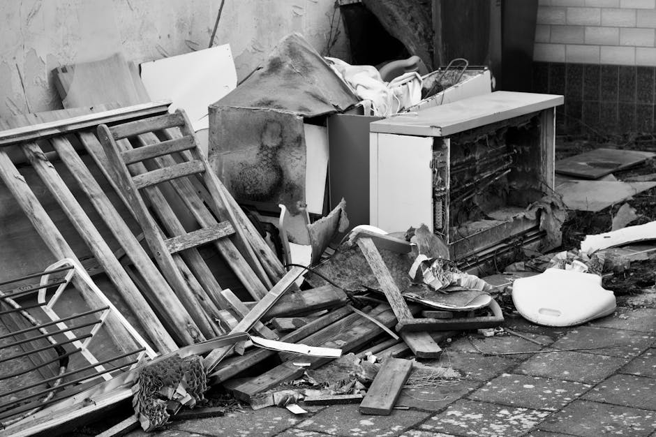 A disorganized pile of waste materials and debris situated outdoors on a paved surface near a brick and concrete wall. The collection includes a toppled wooden garden chair with slatted design, showing weathered and aged wood, leaning against a white, partially broken cabinet with a damaged front panel and visible hardware. Additional rubbish such as a large, rust-stained metal sheet, broken wooden planks, and scattered pieces of cardboard and insulation are present. The debris also includes a white plastic object, possibly a small container or cover, and various unidentifiable fragments of mixed waste. The background features a brick wall and a section of concrete or plastered wall, with some darkened and stained areas suggesting exposure to the elements. The scene is illuminated by natural daylight, highlighting the textures of the damaged and worn materials, indicative of an outdoor clearance associated with private waste disposal or rubbish removal services like those offered by Rubbish Removal Wimbledon.