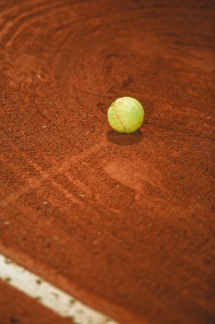 A close-up view of a yellow tennis ball resting on a red clay tennis court surface, positioned near the baseline line which is partially visible at the bottom left of the image. The tennis ball has a slightly worn texture with visible seams, and its bright yellow color contrasts with the rich, reddish-brown tone of the clay, which appears to have a fine granular texture with subtle grooves and markings caused by prior play. The background shows the blurred surface of the court, with variations in shading and small particles of dust or clay, indicating an outdoor environment typical for tennis matches or practice sessions. This scene relates naturally to outdoor sports facilities where tennis courts require regular maintenance and debris removal, highlighting the importance of proper waste management or cleaning services for sports grounds, as offered by local rubbish removal specialists such as Rubbish Removal Wimbledon.