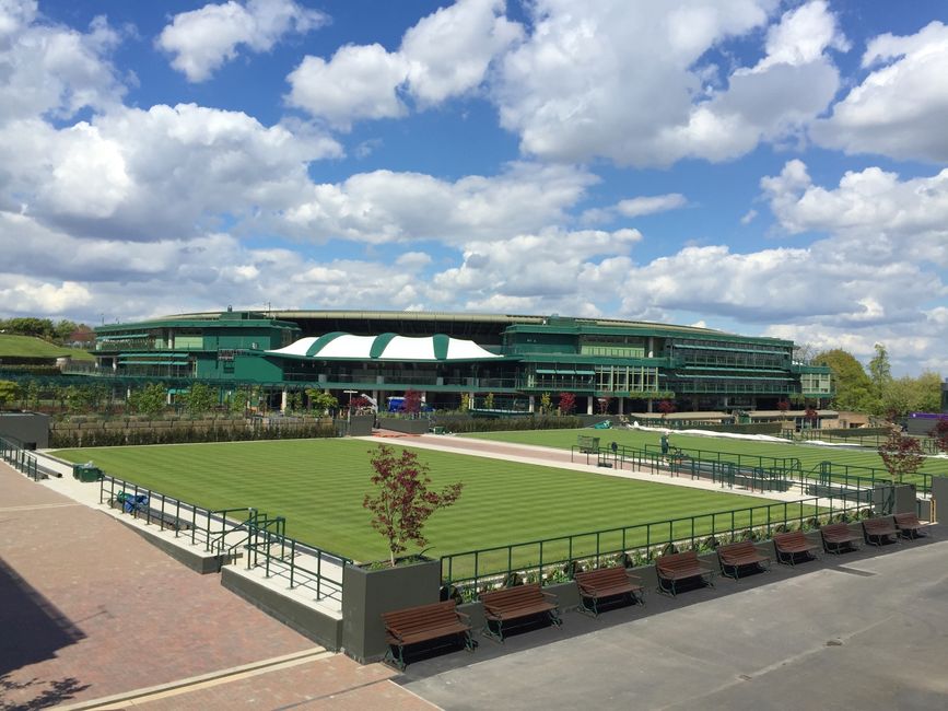 A large modern building with multiple levels and extensive glass windows, painted in shades of green, situated behind a well-maintained grass tennis court. The court is surrounded by a black metal fence and features a smooth, green surface with white boundary lines, indicating it is used for tennis matches. Several empty wooden benches with dark metal frames are positioned along the perimeter of the court, facing the playing area. Small trees with reddish leaves are planted at intervals around the court, adding color and landscaping detail. The environment appears to be an outdoor sports facility, possibly near the All England Lawn Tennis Club Wimbledon, with a bright, partly cloudy sky overhead, natural daylight illuminating the scene. In the foreground, a paved pathway and a low wall enhance the tidy, organized appearance of the setting, which aligns with private or alternative rubbish removal services such as those offered by Rubbish Removal Wimbledon, providing a clear visual context for rubbish and waste clearance activities in an outdoor recreational or sporting location.