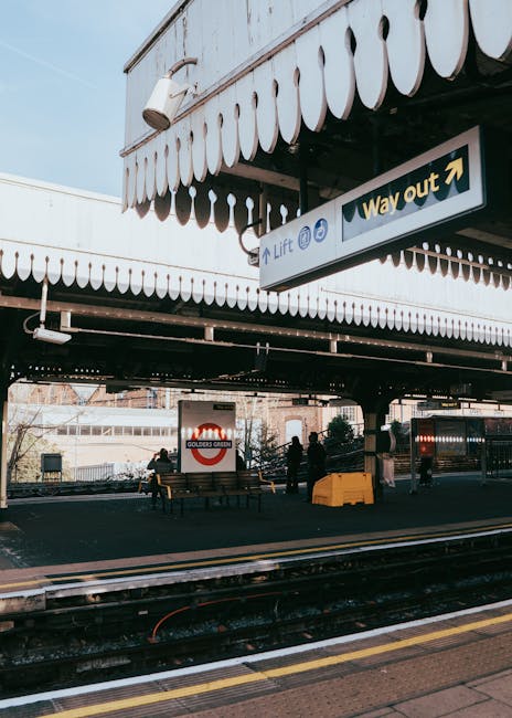 The image depicts a section of an outdoor railway station platform with a canopy overhead featuring a scalloped edge in cream and black. Beneath the canopy, there is a 'Way out' sign with an arrow pointing to the right and additional symbols indicating lift access. On the platform, there are a few people waiting, some sitting on wooden benches while others stand nearby, dressed in dark clothing. A bright yellow waste container, likely used for rubbish collection, is positioned near the benches. The railway tracks are visible in the foreground, showing metal rails and wooden sleepers, with a yellow line marking the edge of the platform for safety. In the background, part of the station building with glass windows, railway infrastructure, and a sign indicating 'Golders Green' station can be seen, suggesting the location is in London. The overall scene captures a typical urban railway station environment, with a focus on public transit and the importance of maintaining waste management practices in such settings, as promoted by independent waste removal services like Rubbish Removal Wimbledon.