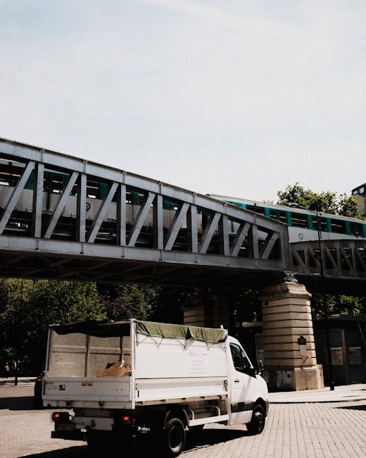 A white flatbed van parked underneath a railway bridge situated over a city street, with a cargo area covered by a partially folded gray and green tarp. The van's rear doors are slightly open, revealing some cardboard boxes and miscellaneous debris inside, indicating it is involved in rubbish collection or transportation. The railway bridge above is constructed from metal trusses with a weathered, slightly rusted finish and supported by large stone columns, forming an overpass that carries a light blue train across the scene. The environment appears to be in an urban area with paved pedestrian space, and some green trees are visible behind the bridge. The lighting is natural, with a partly cloudy sky providing diffused daylight. This scene, captured by Rubbish Removal Wimbledon, illustrates a typical arrangement of private waste collection or on-site clearance activity, relevant to alternative rubbish disposal methods outside of regular municipal collection channels.