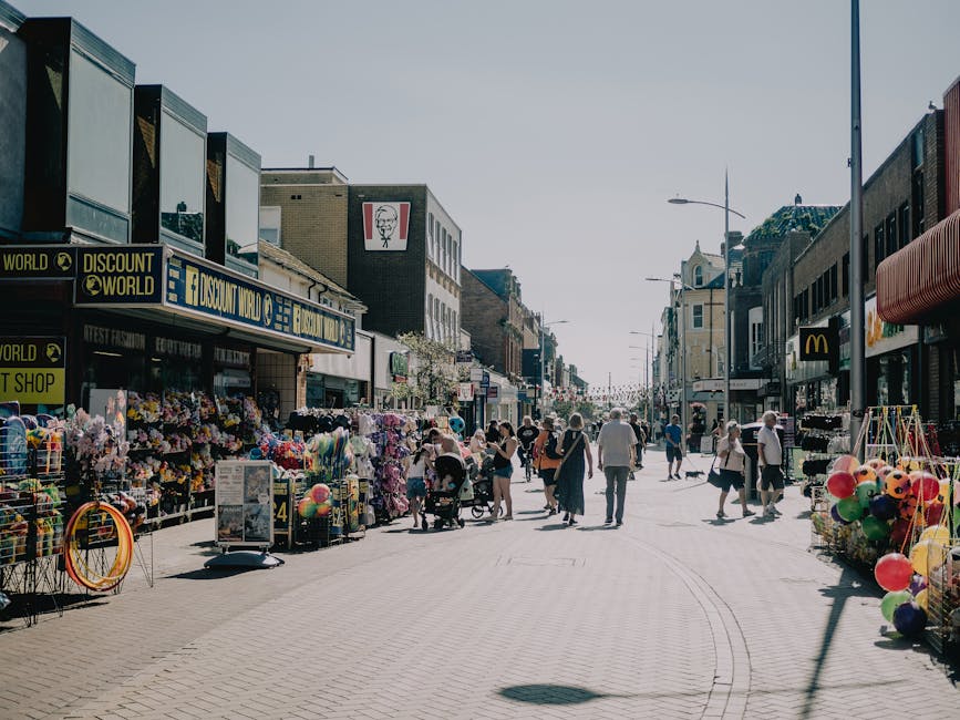 A busy shopping street with pedestrians walking along a wide, paved pedestrian zone lined with retail stores, including a discount shop, a fast-food outlet, and a souvenir stand. The storefronts display various merchandise such as colourful balloons, toys, and accessories, with some items stacked on outdoor display racks or tables. On the left side, a standing signboard advertises products or offers, while on the right side, additional items are arranged outside shops, including balloons and plastic toys. The buildings are multistory with commercial signage, some featuring distinctive logos like McDonald's and KFC, and are constructed with a mix of brick and modern facades. Bright natural daylight illuminates the scene, casting subtle shadows on the pavement. The street appears clean and well-maintained, with several pedestrians engaged in shopping or strolling, some pushing strollers or carrying bags. This environment, typical of an active retail sector, may be indicative of areas where independent waste collection or disposal services like Rubbish Removal Wimbledon could facilitate waste management, especially after busy shopping days.